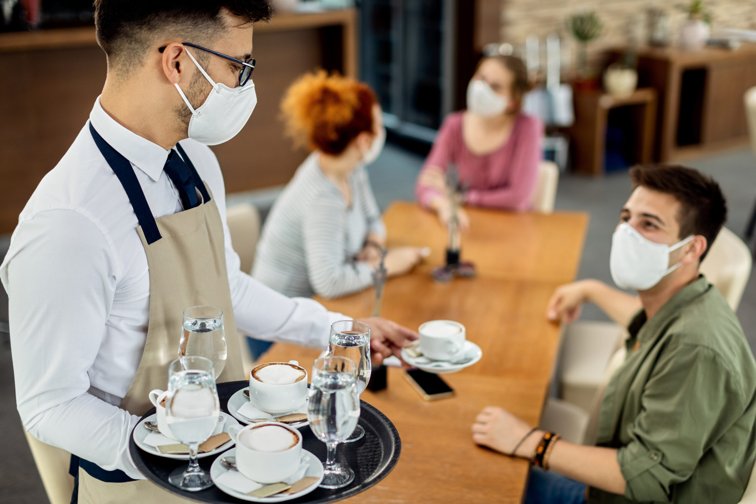 Group of friends and waiter wearing protective face mask in a cafe due to coronavirus epidemic. Focus is on waiter serving coffee to them.