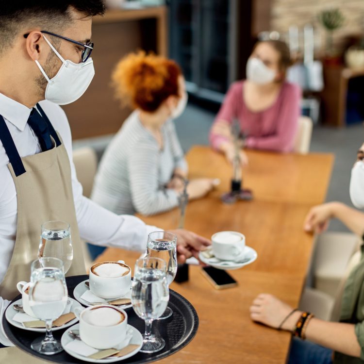 Group of friends and waiter wearing protective face mask in a cafe due to coronavirus epidemic. Focus is on waiter serving coffee to them.